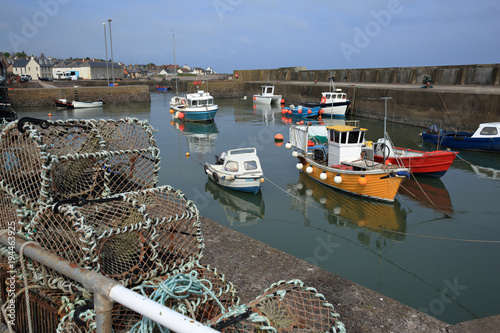 The still tranquil harbour at Johnshaven in north east Scotland UK. Small yellow red and green fishing boats float in a harbour surrounded by a concrete sea wall. Lobster pots stored by fishing boats