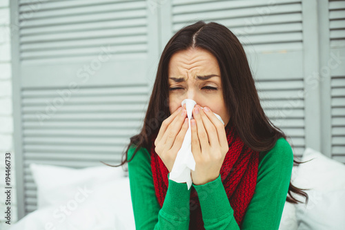 Close up portrait of a young sick woman with a runny nose in a red scarf lying on the bed at home and treated. The concept of health and disease.