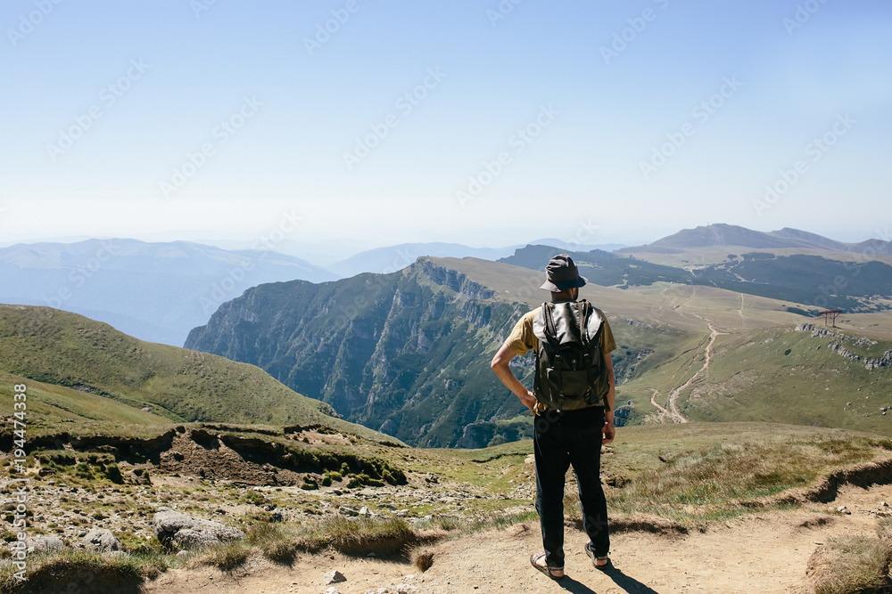 Young hipser man with backpack enjoying mountain view on bright sunny day. Hiker on mountain background. Empty cloudless sky. Young hipser man with backpack enjoying mountain view on bright sunny day. Hiker on mountain background. Empty cloudless sky.