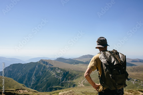 Young hipser man with backpack enjoying mountain view on bright sunny day. Hiker on mountain background. Empty cloudless sky.