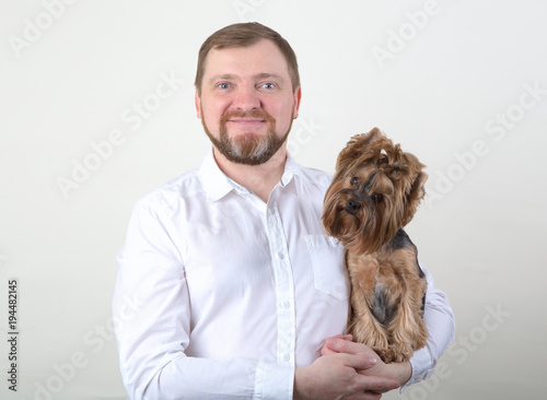 Bearded man holds a small Yorkshire terrier dog in his arms on the gray background.
