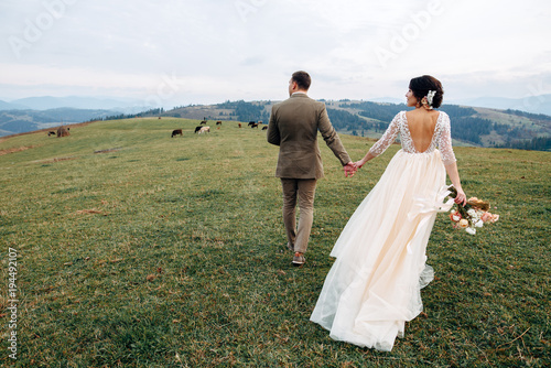 The groom and bride in beige dress with naked back are walking on green hill with mountains on background