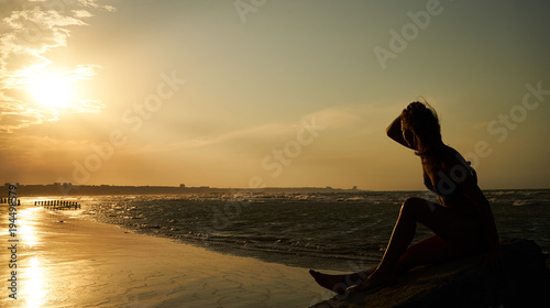 Happy carefree woman enjoying beautiful sunset on the beach. Profile of a young woman silhouette in swimsuit sitting and watching on the sun. Vacation travel holidays and summer time concept