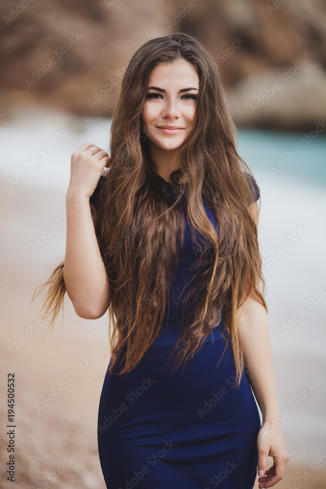 Pretty young smiling girl in a tight dark blue dress dreams on the beach. Blurred background. Vertical orientation.
