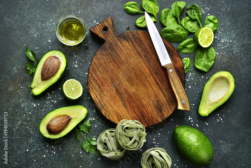 Uncooked spinach pasta (tagliatelle) and green vegetables (spinach, lime, avocado) with empty wooden cutting board.Top view with copy space .