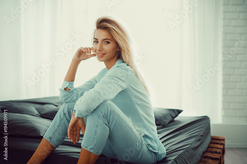 Natural pretty blonde girl in casual clothes sitting on bed in bright bedroom photo