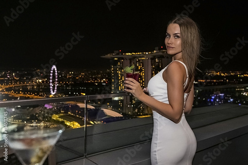 красивая молодая женщина в белом платье с глубоким декольте и большой грудью сидит в баре на крыше на фоне огней ночного сингапур