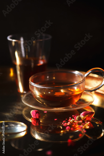 Vertical close-up shot of small glass teapot and cup with hot red tea, dried rose petals, pocket magnifier on golden chain on golden tray. Evening light. Golden color concept.