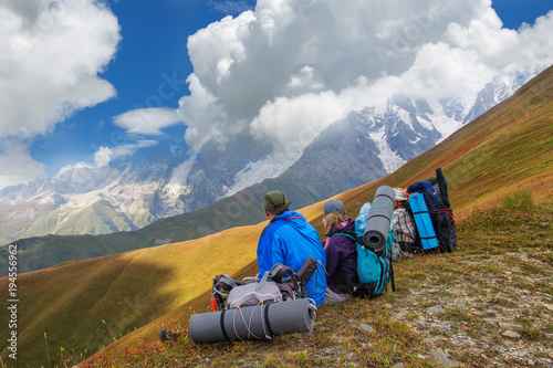 A group of tourists with big backpacks relaxes on a green meadow with mountain views
