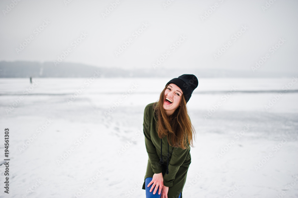 Funny girl wear on long green sweatshirt, jeans and black headwear, at frozen lake in winter day.