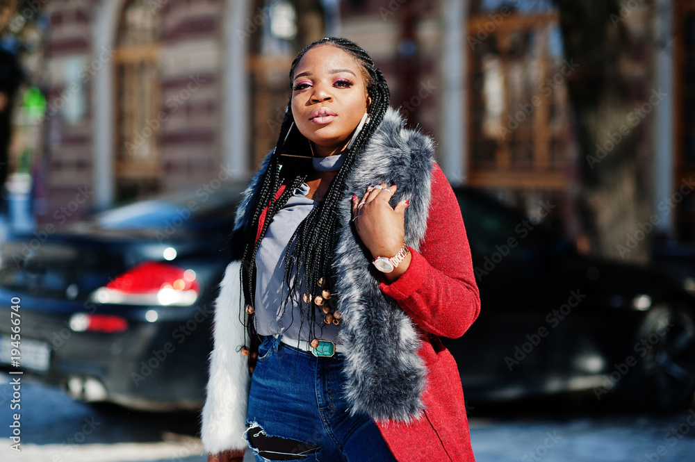 Fototapeta premium Rich african american girl in red coat and fur on winter street at sunny weather against black business car. Black stylish woman.