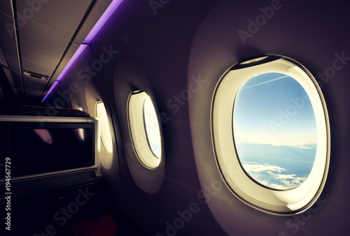 Stunning wide angle view of three airplane windows seen from a business class seat on a long haul widebody aircraft. Big TV screen on the left. Purple mood lighting on the ceiling. Bright day outside.