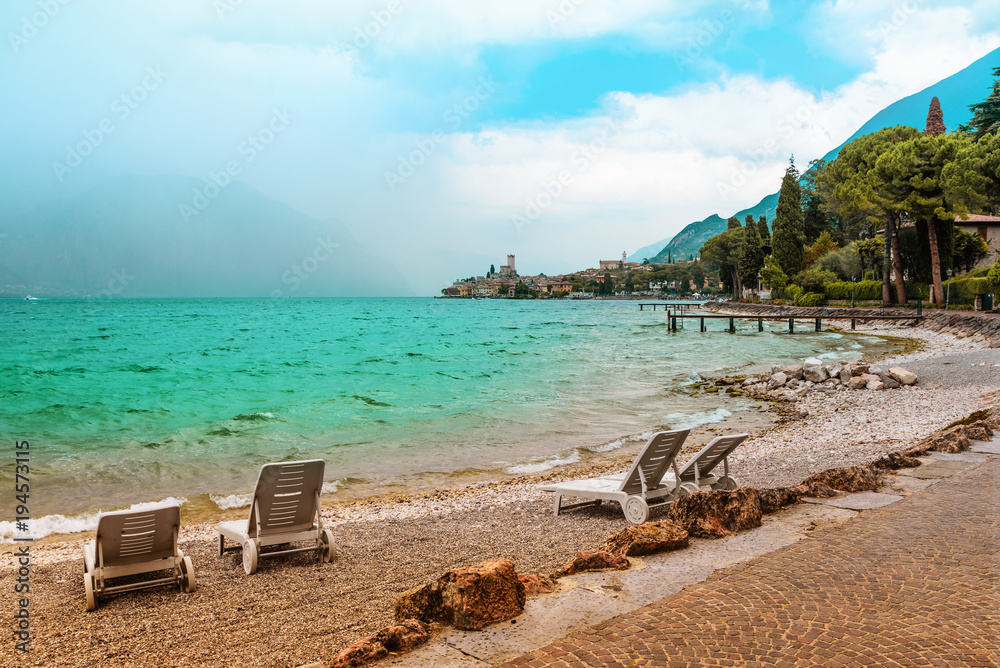 Liegestühle am Gardasee Badestrand Malcesine, türkisblaues Wasser und Blick zur Burg Liegestühle am Gardasee Badestrand Malcesine, türkisblaues Wasser und Blick zur Burg
