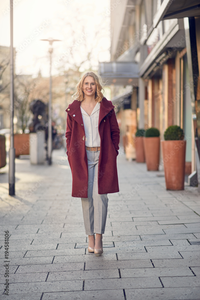 Attractive stylish young woman in a maroon overcoat standing with her hands in her pockets in a city street smiling at the camera