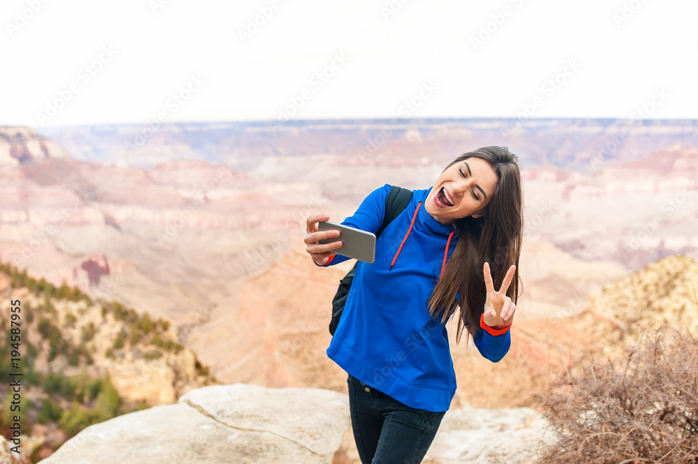 Young beautiful girl at Grand Canyon National Park viewpoint, Arizona, USA  with a smartphone taking selfportrait and making the peace sign with hand.
