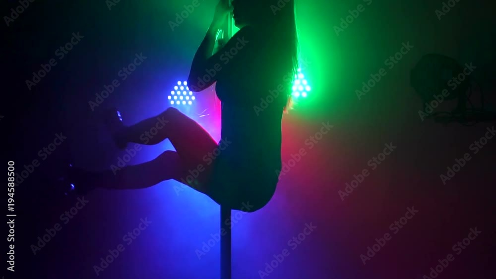 A flexible dancer performs a complex exercise on a pylon in the dark with the light of green, red and blue projectors. Pole dance. Slow motion. Silhouette. A flexible dancer performs a complex exercise on a pylon in the dark with the light of green, red and blue projectors. Pole dance. Slow motion. Silhouette.