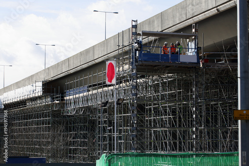 Motorway maintenance as three construction workers in orange jackets stand high up on a large scaffold structure inspecting a section of highway bridge/flyover. Civil engineering transport project.