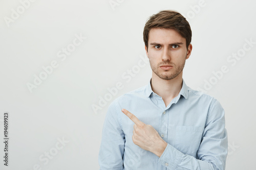 Employee do not believe strange customer, looking at him suspiciously. Portrait of handsome caucasian man in formal shirt pointing left with index finger, being doubtful and not sure in person