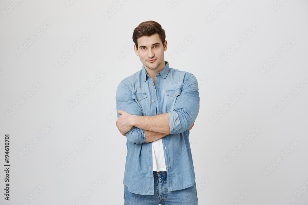 Fototapeta premium Attractive young sportsman with beautiful eyes, confident expression and crossed hands near white background. Artist just finished his painting, looking with satisfaction, enjoying result