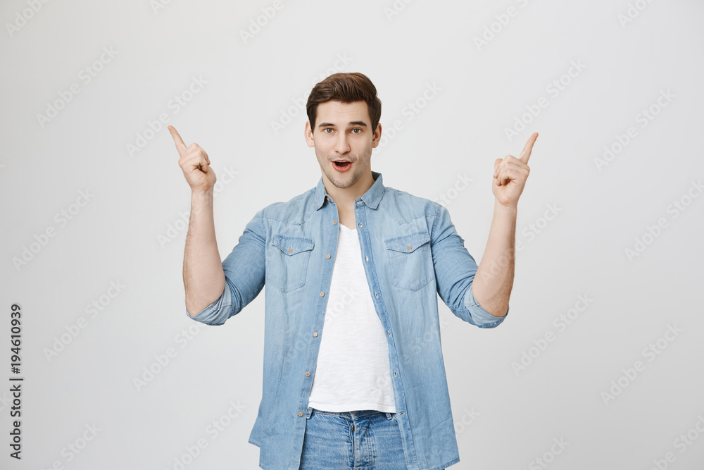 Excited dark-haired guy in denim shirt sees something unbelievable, indicates with fore finger at copy space for advertisement or promotional text. Amazed young european stylish male points upwards Excited dark-haired guy in denim shirt sees something unbelievable, indicates with fore finger at copy space for advertisement or promotional text. Amazed young european stylish male points upwards