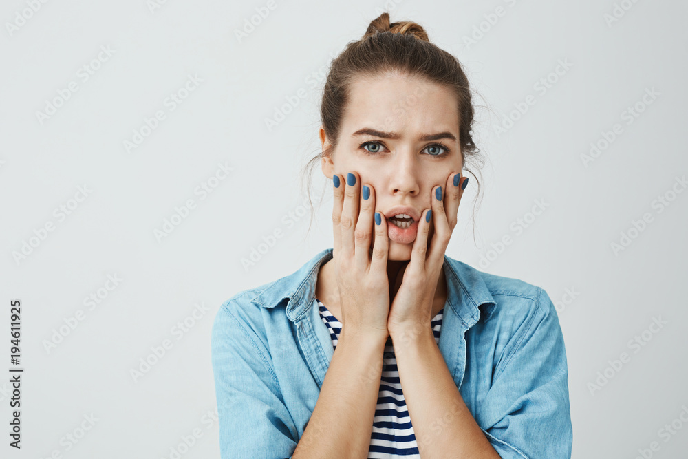 Girl is worried and shocked. Studio portrait of beautiful adult woman with bun hairstyle holding palms on face and looking with nervous and troubled expression, standing with opened mouth.