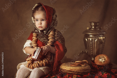 Fotografija A small sweet girl in a national costume sits on a wooden table near the samovar and eats pancakes