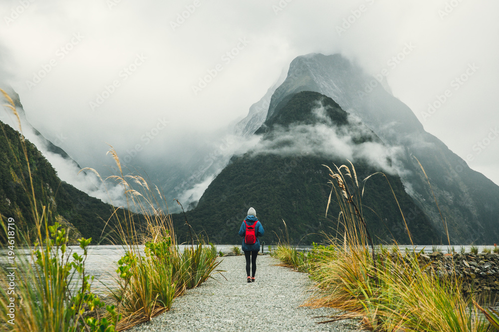 One woman stands in front of mountains in blue jacket with red backpack