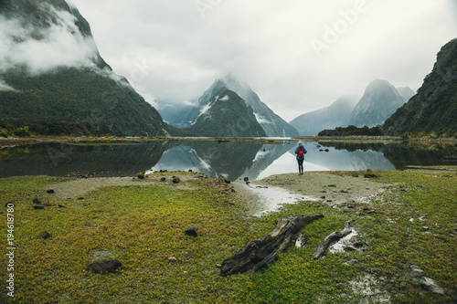 One woman stands in front of a lake mirroring mountains in blue jacket with red backpack