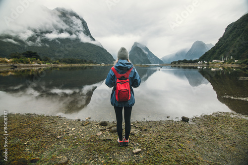 One woman stands in front of a lake mirroring mountains in blue jacket with red backpack