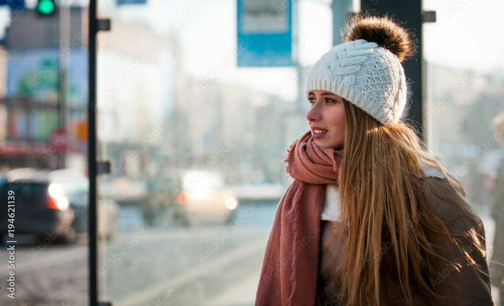 Beautiful woman in wool hat waiting at bus stop during winter day