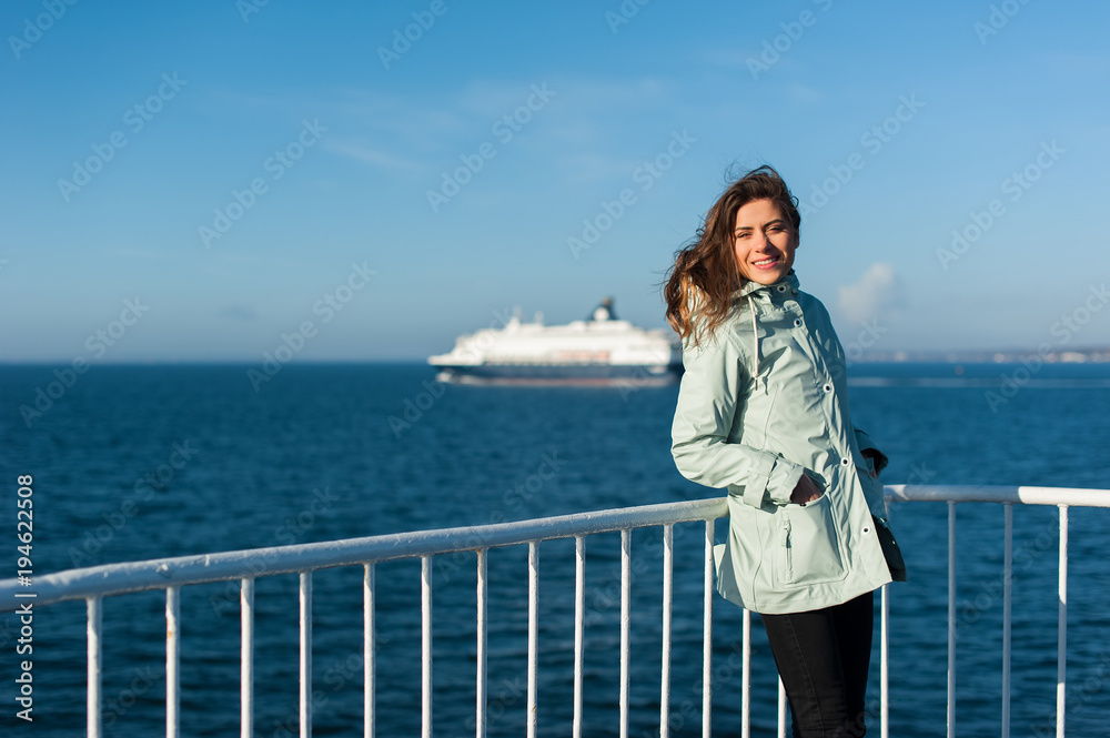 Young traveller woman looking at camera and smiling, on the sea sailing a ferry, with big boat cruise liner or ferry on the background, wearing a rain jacket.
