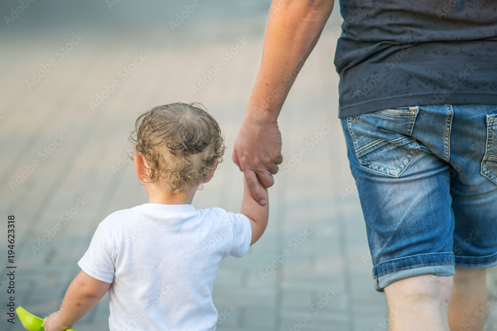 A young father holding his little baby son hand. A parent and a boy walking together in summer park. Outdoor, day, kid, dad, walk, family