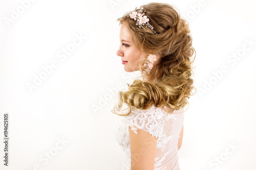 young blonde girl in a white wedding dress and jewelry in hairstyle with big curls smiling like angel on a white wall background portrait