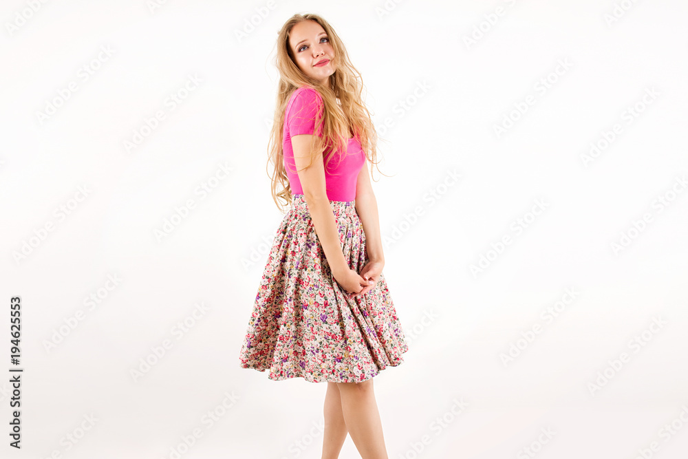 young cutest blonde girl in a pink combination and skirt with flowers with long hair curls  standing  and smiling  like a barbie hesitates on a white wall background