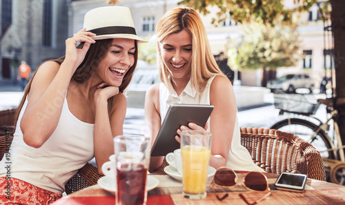 Friends meeting in a cafe. Young women drinking coffee and have fun with tablet. Consumerism, lifestyle concept