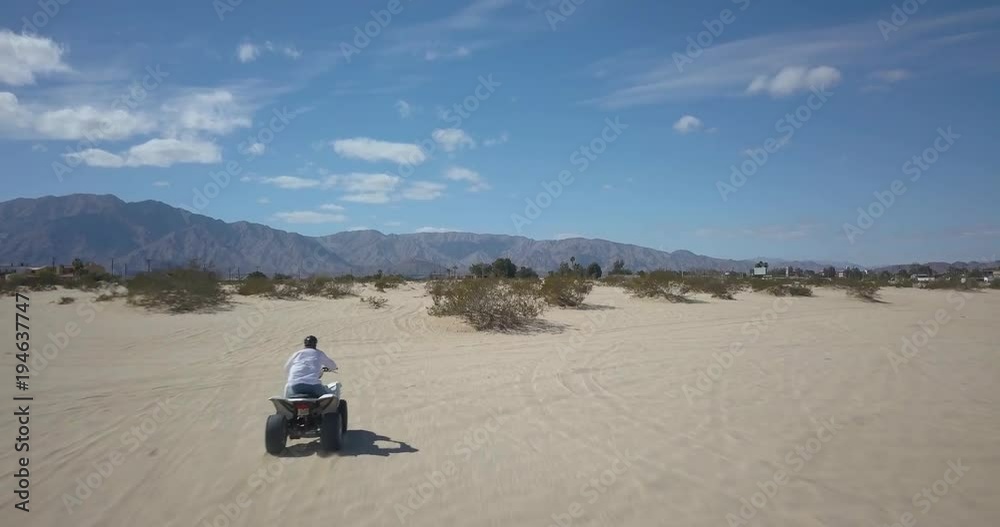 Aerial camera tracking and flying over the head of an ATV rider moving rapidly through sand and scrub vegetation toward mountains in the distance. Aerial camera tracking and flying over the head of an ATV rider moving rapidly through sand and scrub vegetation toward mountains in the distance.