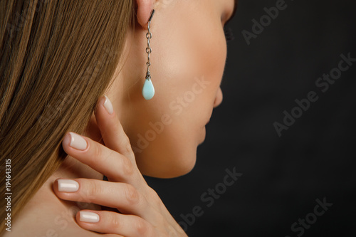 красивая молодая женщина стоит спиной к камере и положила пальцы руки на шею, а в ухе у нее красивая сережка с голубым камнем 