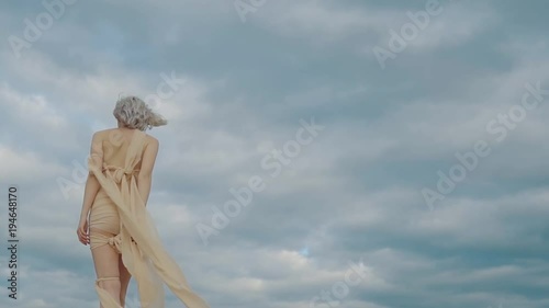 Half-naked goddess is strolling against cloudy sky. Her clothes is waving, back view of graceful womans body