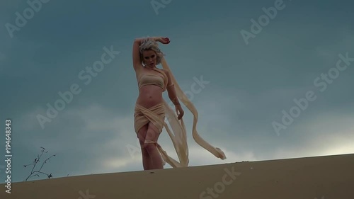 Sexy young blonde is posing outdoors on a sand in background of rainy sky. She is touching scraps of her dress swaying by wind, abstract and fashionable shot