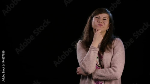 An attractive young lady wearing a pink sweater standing against a black background. The lady supports her chin showing hesitation and dissatisfaction . Medium shot