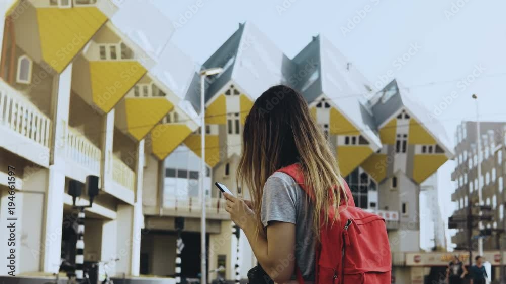 Caucasian girl with phone near amazing buildings. Traveler takes photo of Cube House, Rotterdam, walks forward. 4K.