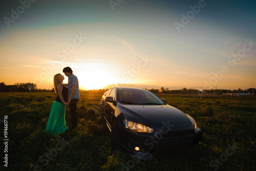 A loving couple watching beautiful bright romantic sunset after journey, standing leaning against blue sport car. The fields around them. The young man shows to his girlfriend evening sun © ANR Production