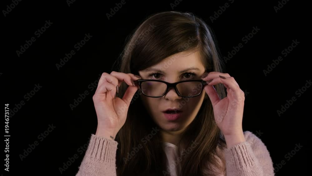 An attractive young lady with with glasses wearing a pink sweater stares in astonishment against a black background. Close-up