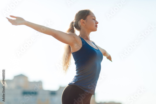 Fototapeta Naklejka Na Ścianę i Meble -  Young fitness woman in a blue shirt and leggings smile spread her hands to the sky for success in waiting the sun shines in her face, in stadium on the background of houses.