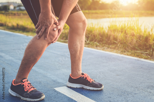 Injury from workout concept : Asian man use hands hold on his knee while running on road in the park. Shot in morning time, sunlight and warm effect with copy space for text or design