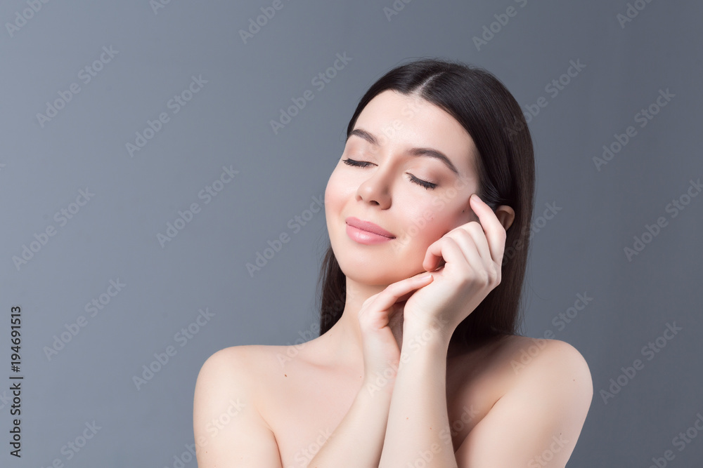 Beautiful young woman with clean skin isolated on grey background. Portrait of beauty girl with natural nude makeup touching her face. Spa, skincare and wellness concept close up. Beautiful young woman with clean skin isolated on grey background. Portrait of beauty girl with natural nude makeup touching her face. Spa, skincare and wellness concept close up.