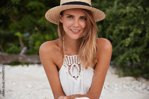 Portrait of gorgeous relaxed female model wears summer straw hat and white dress, has good rest alone outdoor, breathes fresh air and admires sunshine. People, season and recreation concept.