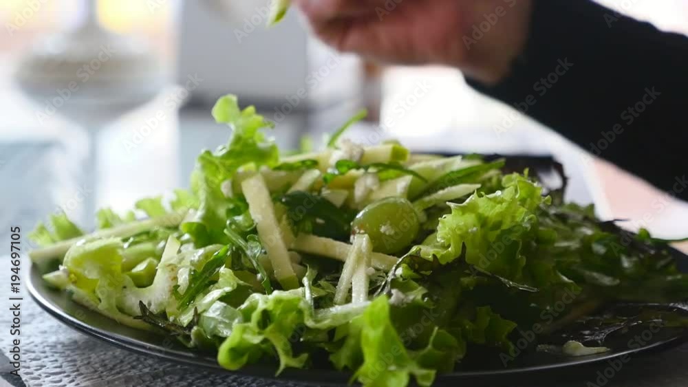 A closeup of a flat plate full of fresh green salad. Some hand uses a metallic fork to take it. The windows are in the background. A closeup of a flat plate full of fresh green salad. Some hand uses a metallic fork to take it. The windows are in the background.