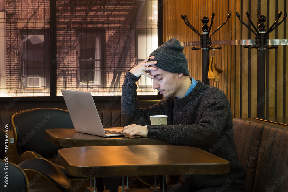 Tired exhausted man holds hand on forehead at cafe. Young male person with closed eyes in cafe in front of laptop computer