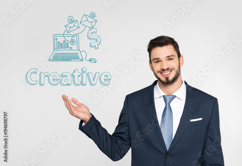 handsome young businessman showing leadership icons and smiling at camera isolated on grey